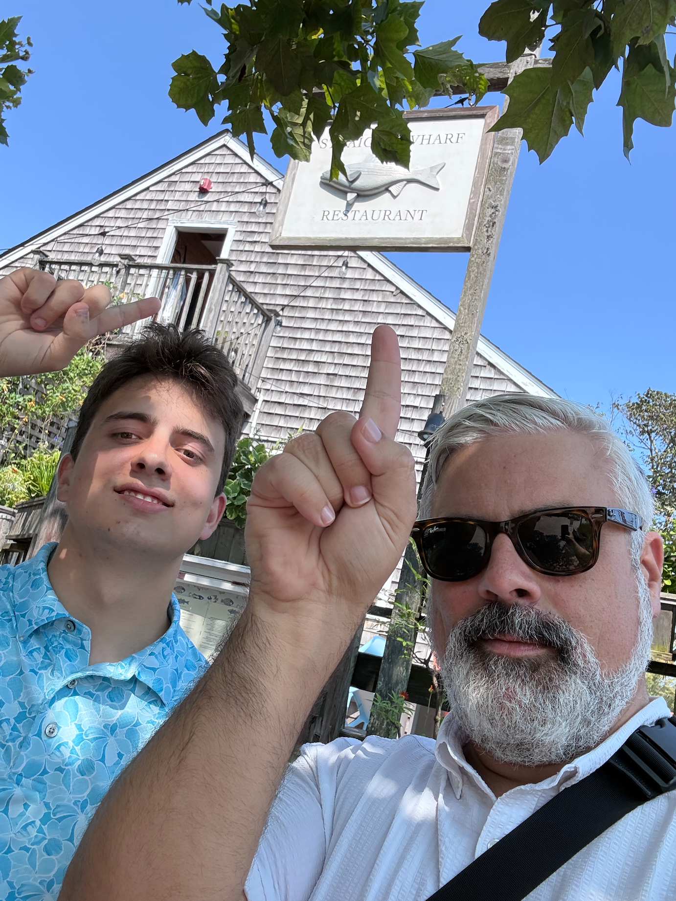 Two people raising fingers in front of a white shingled house on a sunny day.
