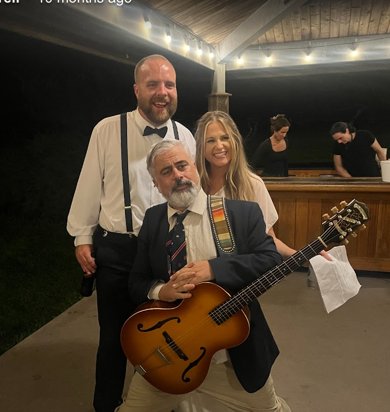 Three people pose at a nighttime event; one holds a guitar in front of a lit bar.