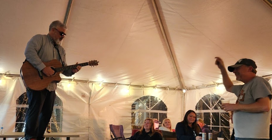 Two men performing music under a lit white tent, one playing guitar and one singing into a microphone.