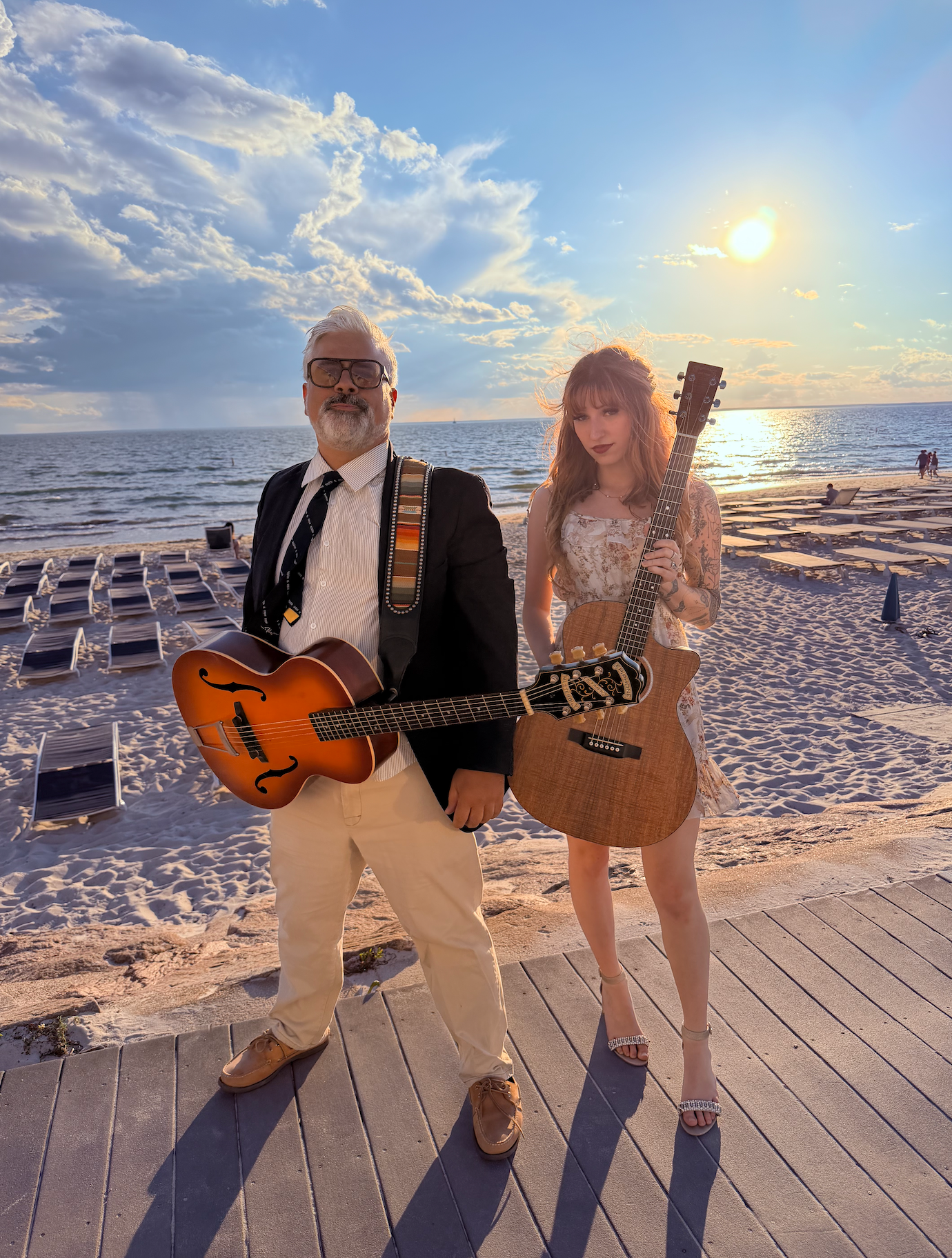Two people holding guitars stand on a boardwalk by the ocean at sunset.