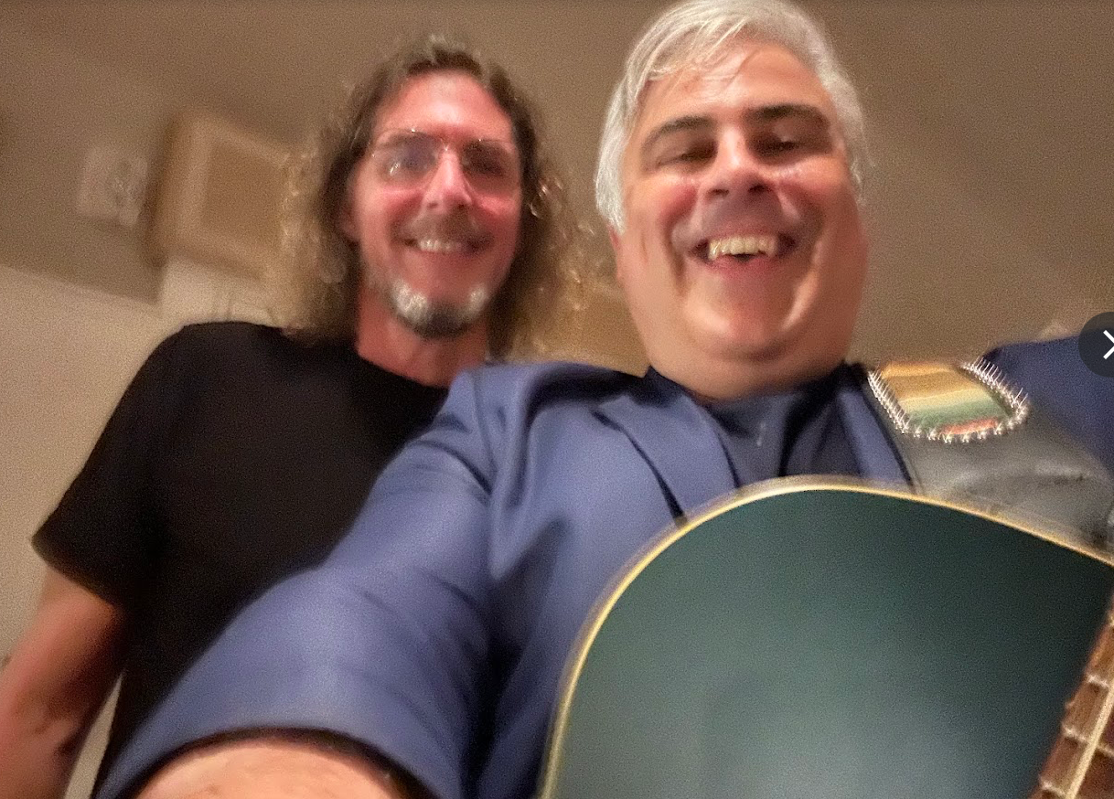 Two smiling people indoors; one holds a guitar in the foreground.