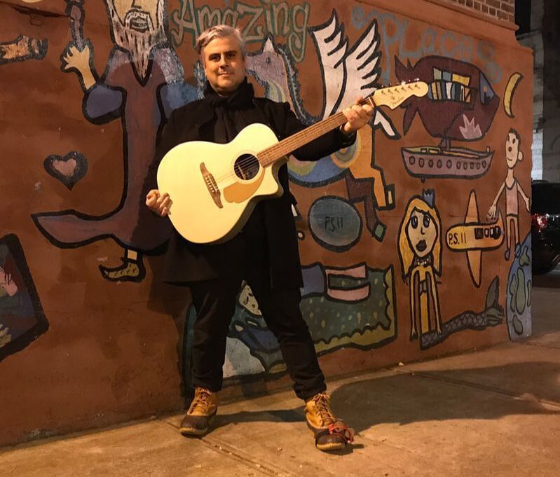 Person holding a guitar, standing against a colorful mural at night.