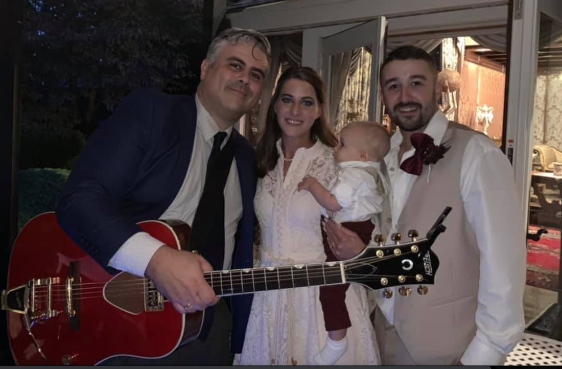 A man is holding a guitar in front of a bride and groom and a baby.