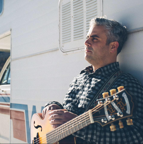 Man leaning against an RV, holding a guitar and looking off to the side.