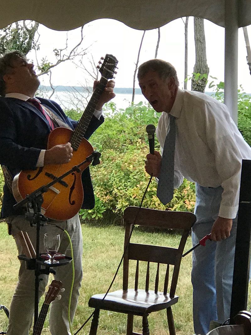 Two musicians under a canopy, one holding a guitar and one at a microphone beside a chair.