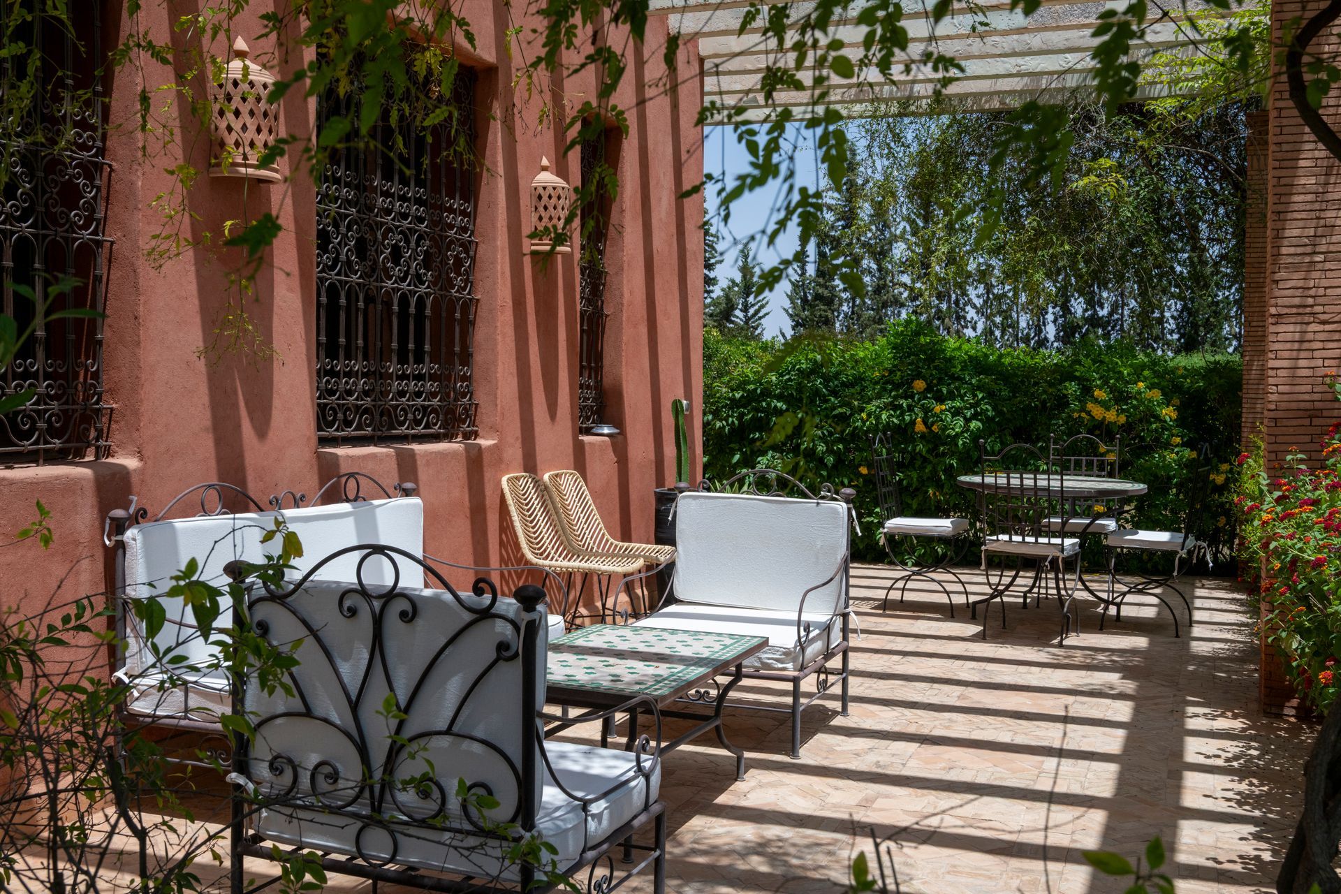 Patio with wrought-iron chairs and tables beside a pink stucco building, shaded by vines and sunlight.