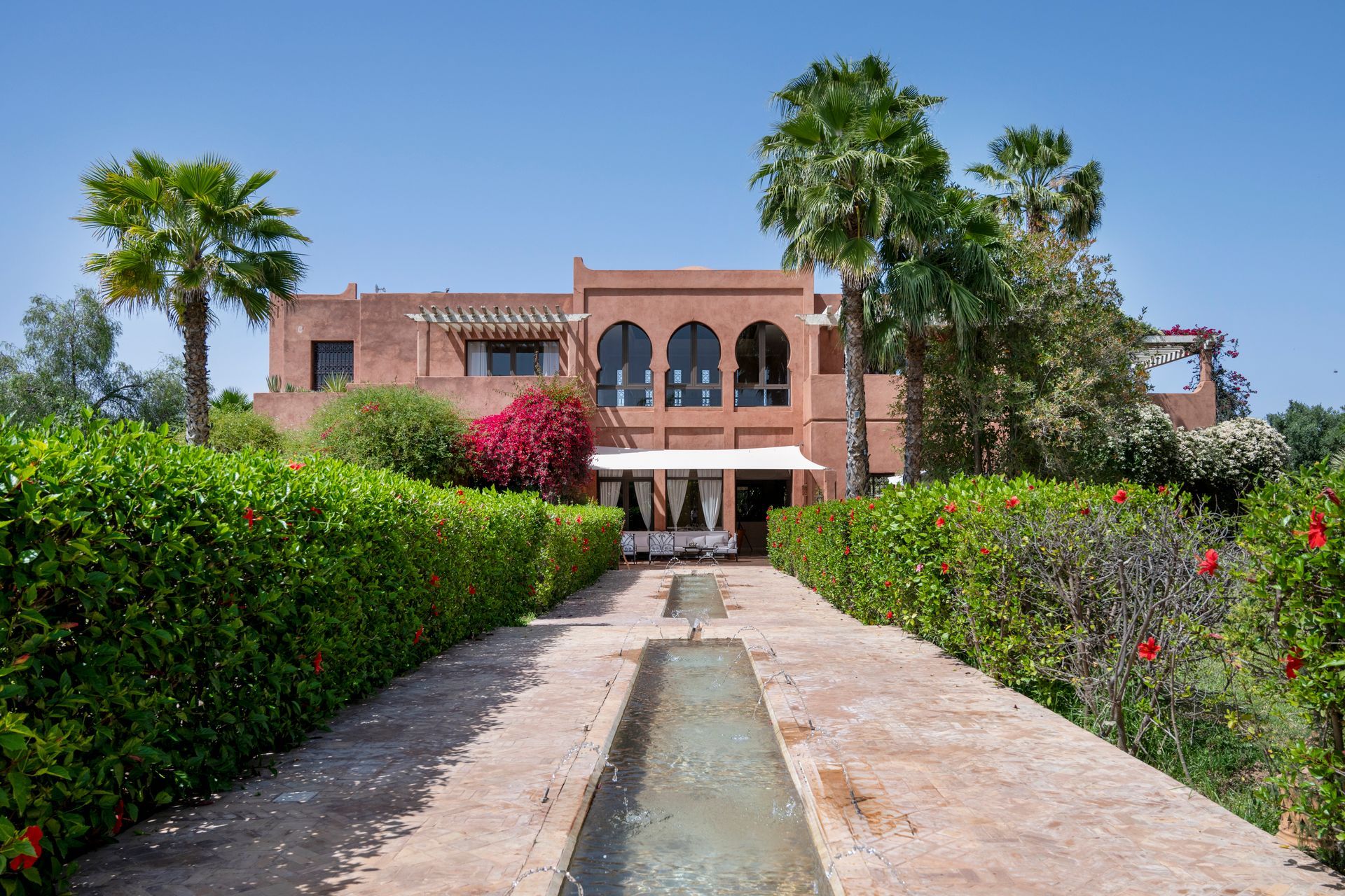 Brick villa at end of a hedge-lined path, with palm trees and a narrow water channel in front