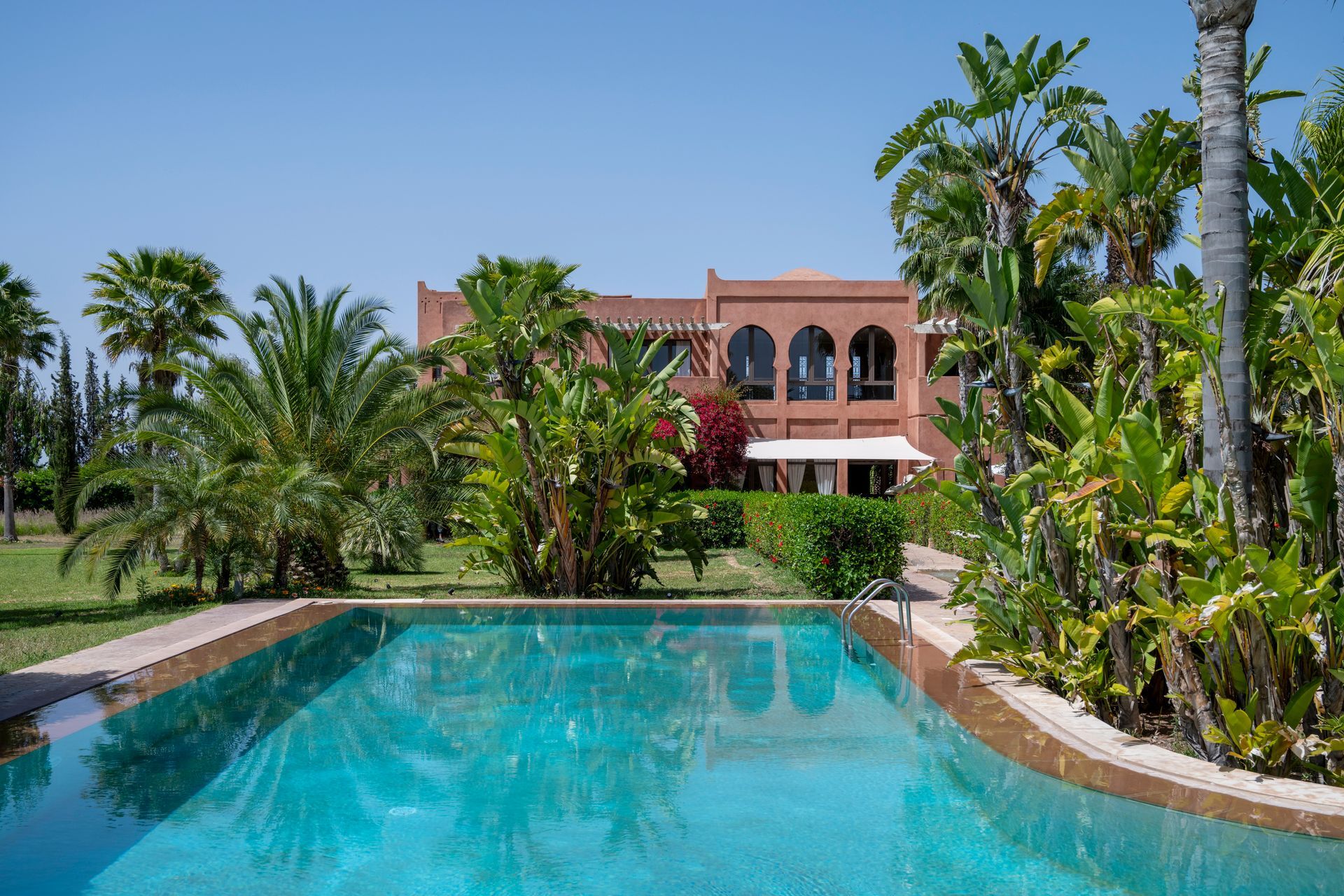 Red-brick building with arched windows, palm trees, and lush green garden under a clear blue sky