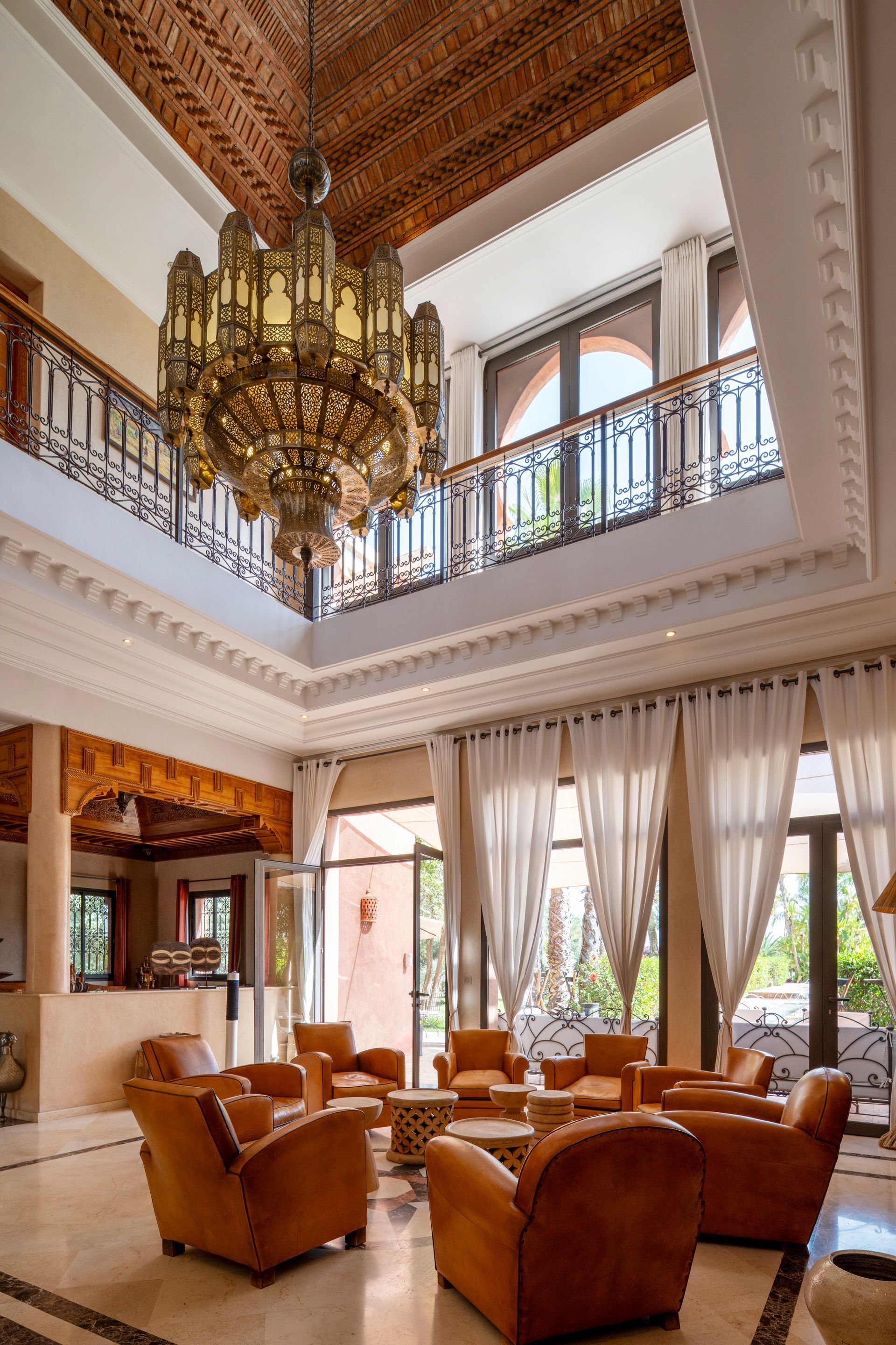 Luxurious living room with leather armchairs, tall windows, and a large chandelier under a carved wood ceiling