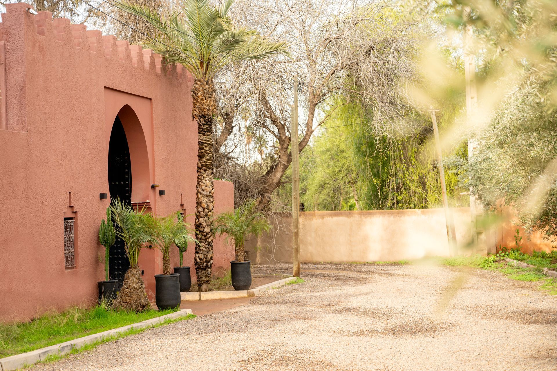 Sunlit courtyard with terracotta building, arched doorway, potted plants, and gravel path