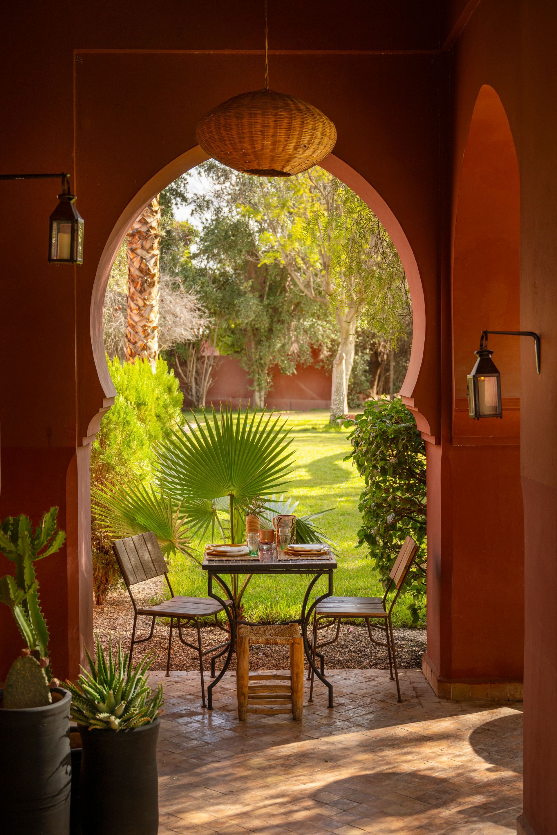 Cozy patio with arched doorway, wicker table and chairs, potted plants, and a sunlit garden beyond