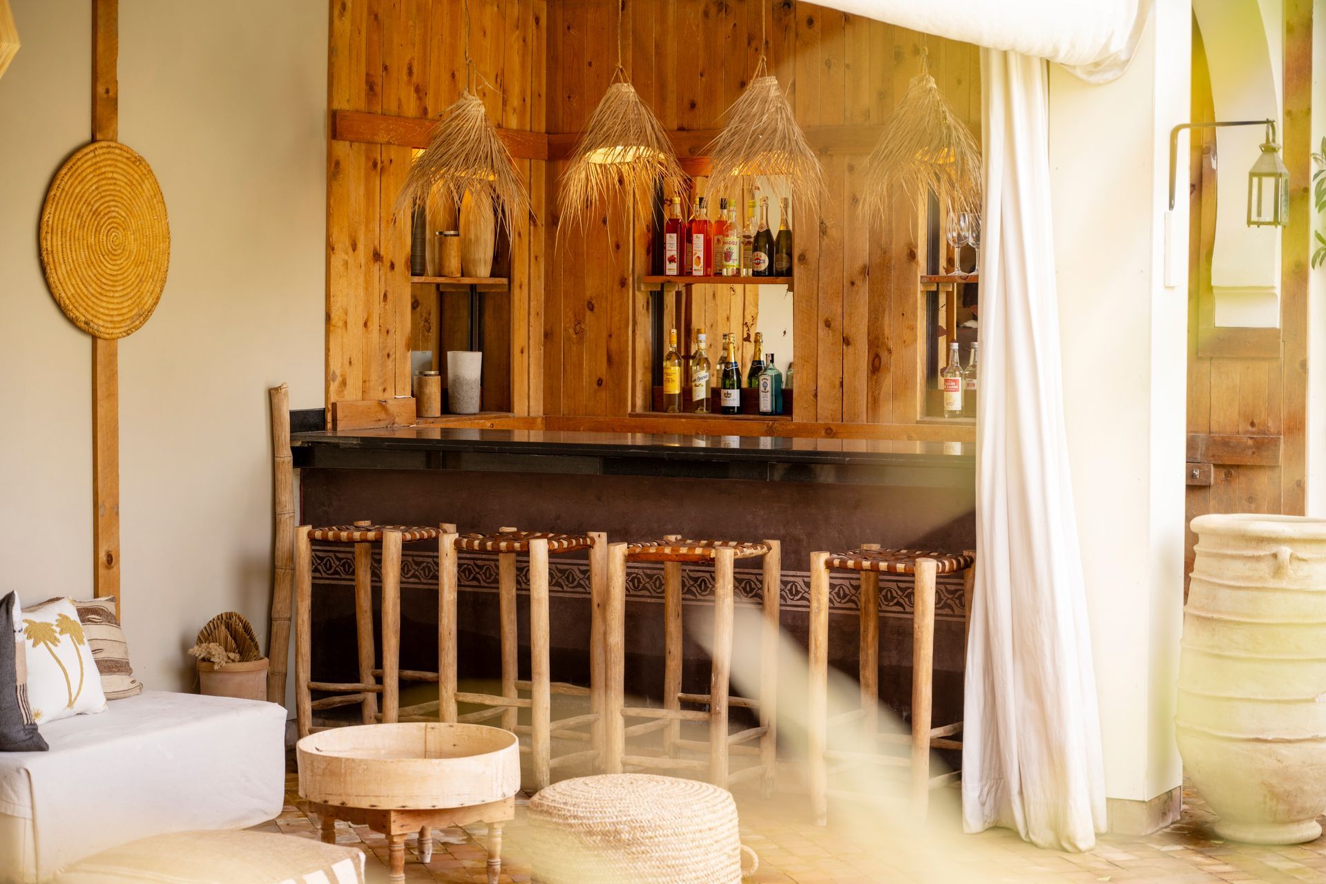 Cozy wooden bar with stools, shelves of bottles, and sheer curtains in warm sunlight