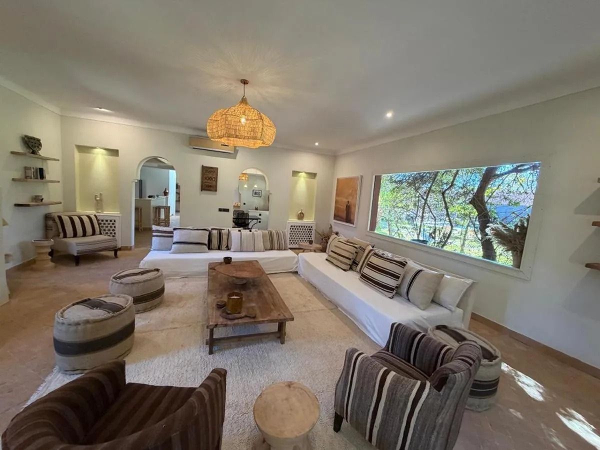 Bright living room with curved sectional sofas, patterned chairs, low coffee table, and large window view of trees