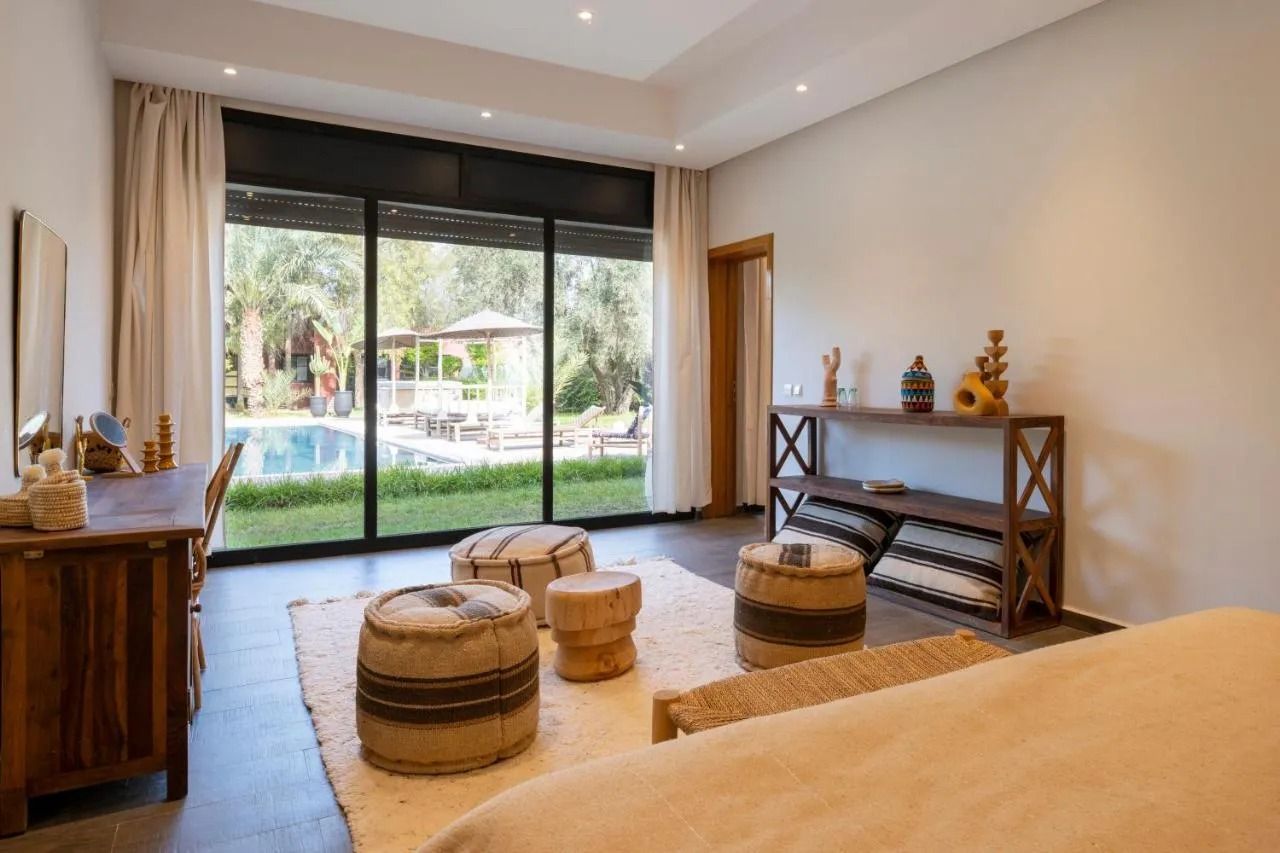 Bright modern bedroom with a large sliding glass door, woven stools, rug, and console table facing a garden view