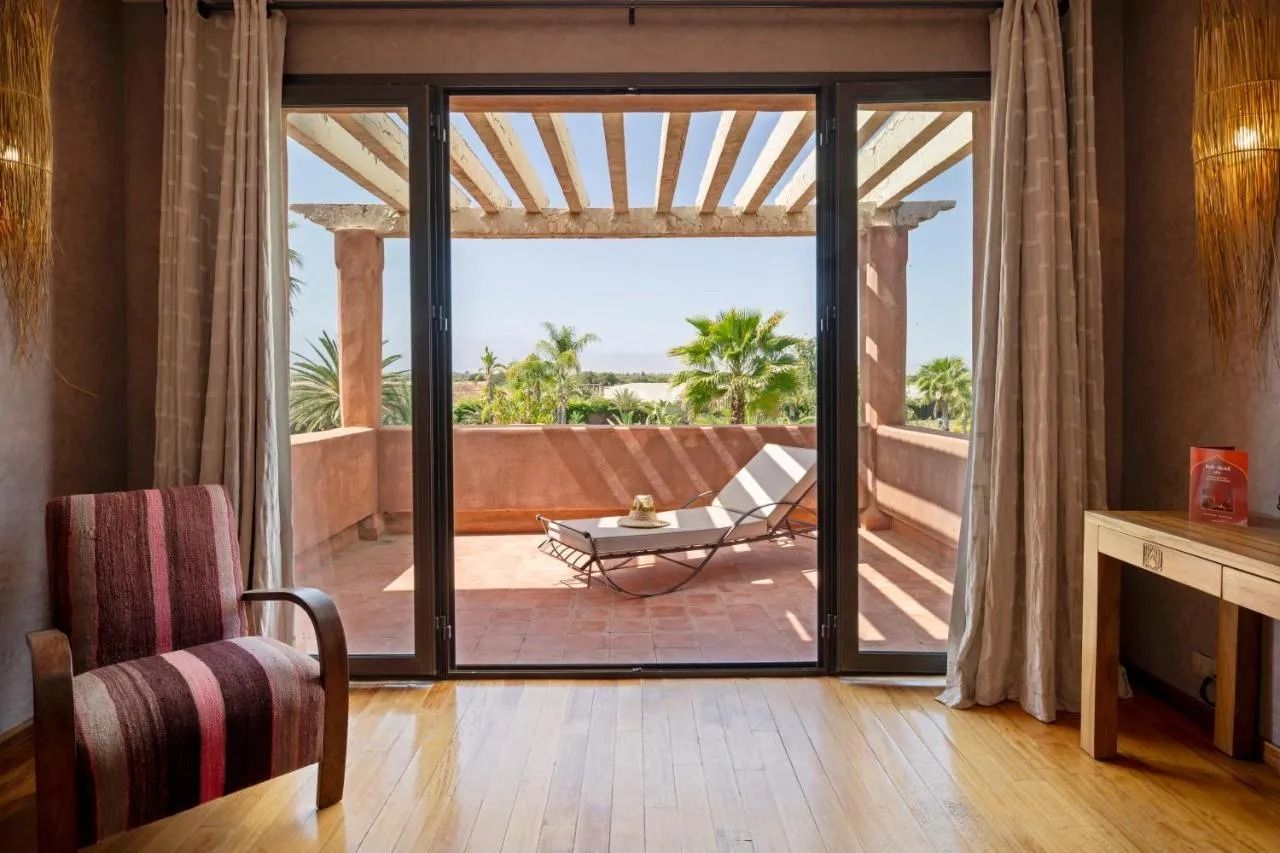 Bright bedroom with open balcony doors, two lounge chairs, and a sunlit ocean view.