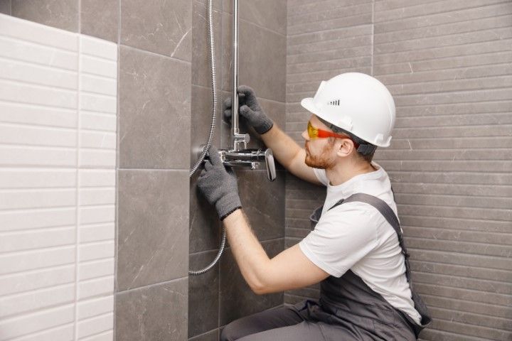 Worker installing a wall-mounted fixture in a tiled bathroom, wearing a hard hat, gloves, and safety glasses.