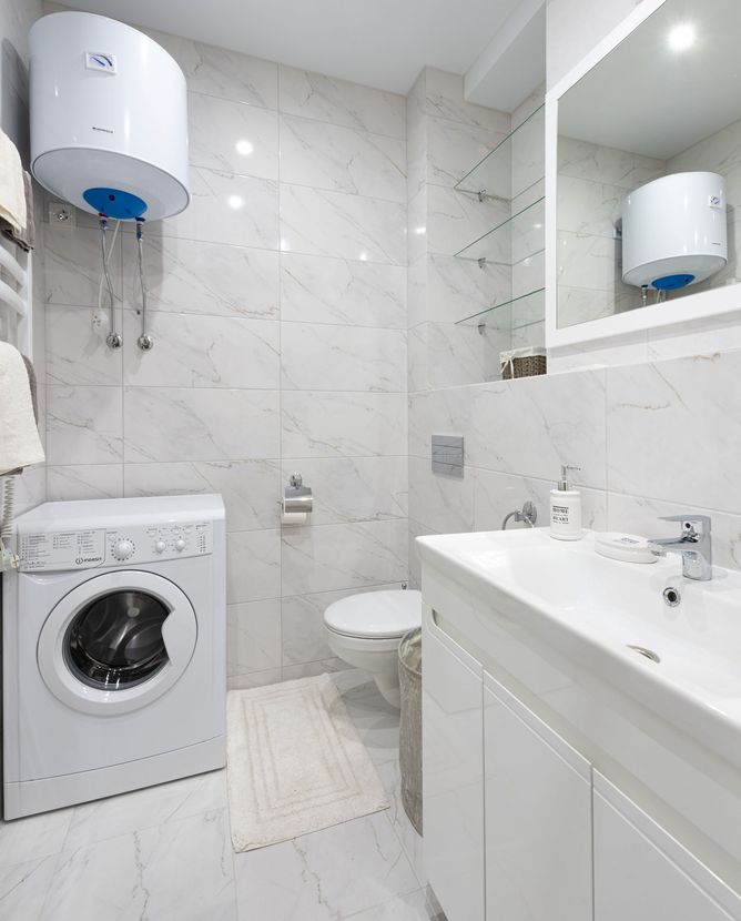 Small white bathroom with washing machine, toilet, sink vanity, mirror, and wall-mounted water heater.