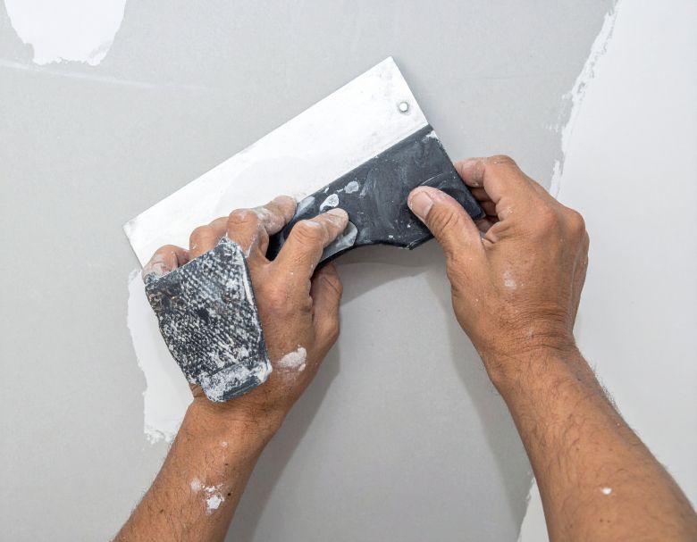 Hands smoothing drywall compound with a putty knife and sanding sponge on a gray wall.