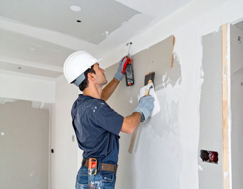 Construction worker in hard hat, applying drywall compound to a wall with a trowel. Interior setting.