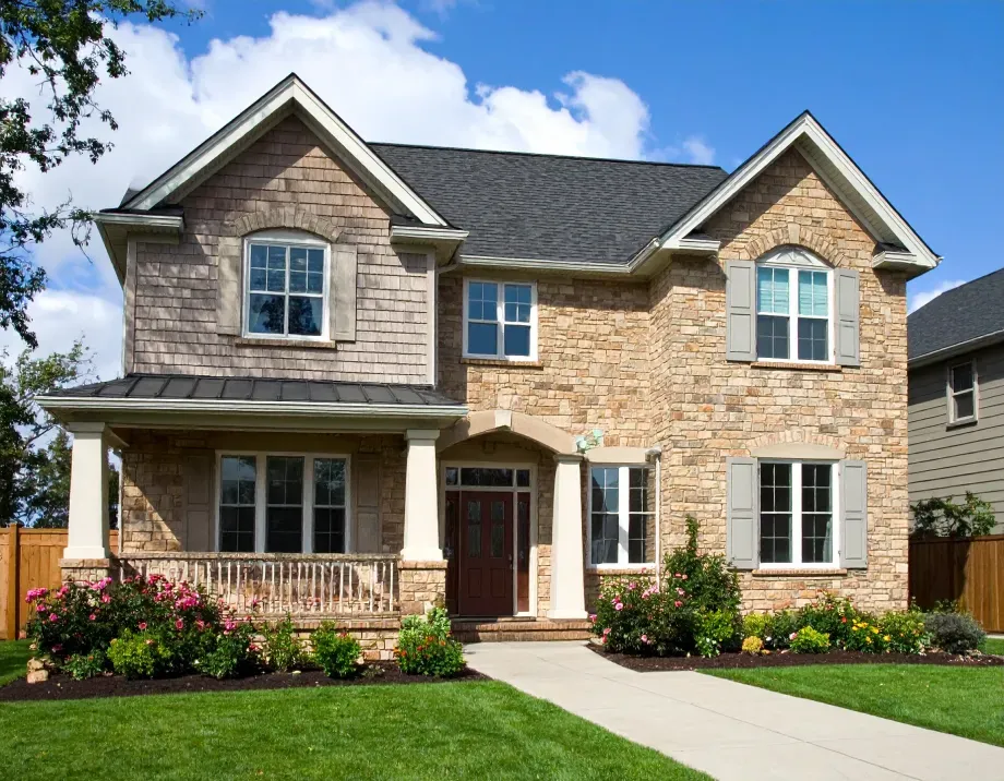 Two-story brick house with a porch, gray roof, and manicured lawn on a sunny day.