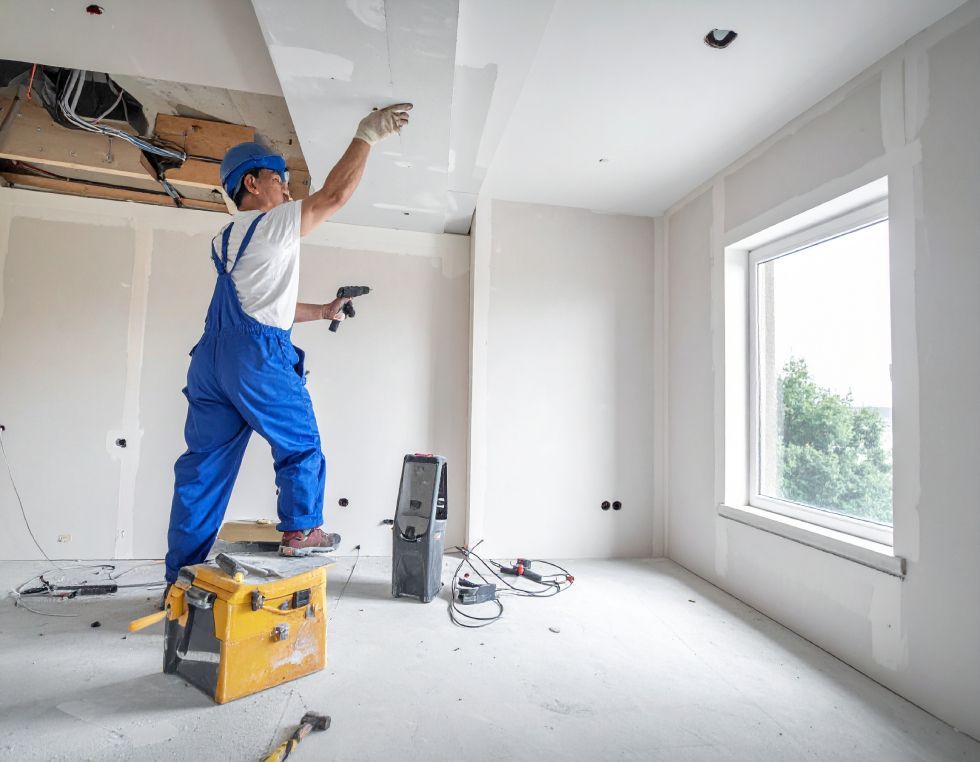 Construction worker in blue coveralls and hard hat, working on a ceiling in a room with a window.