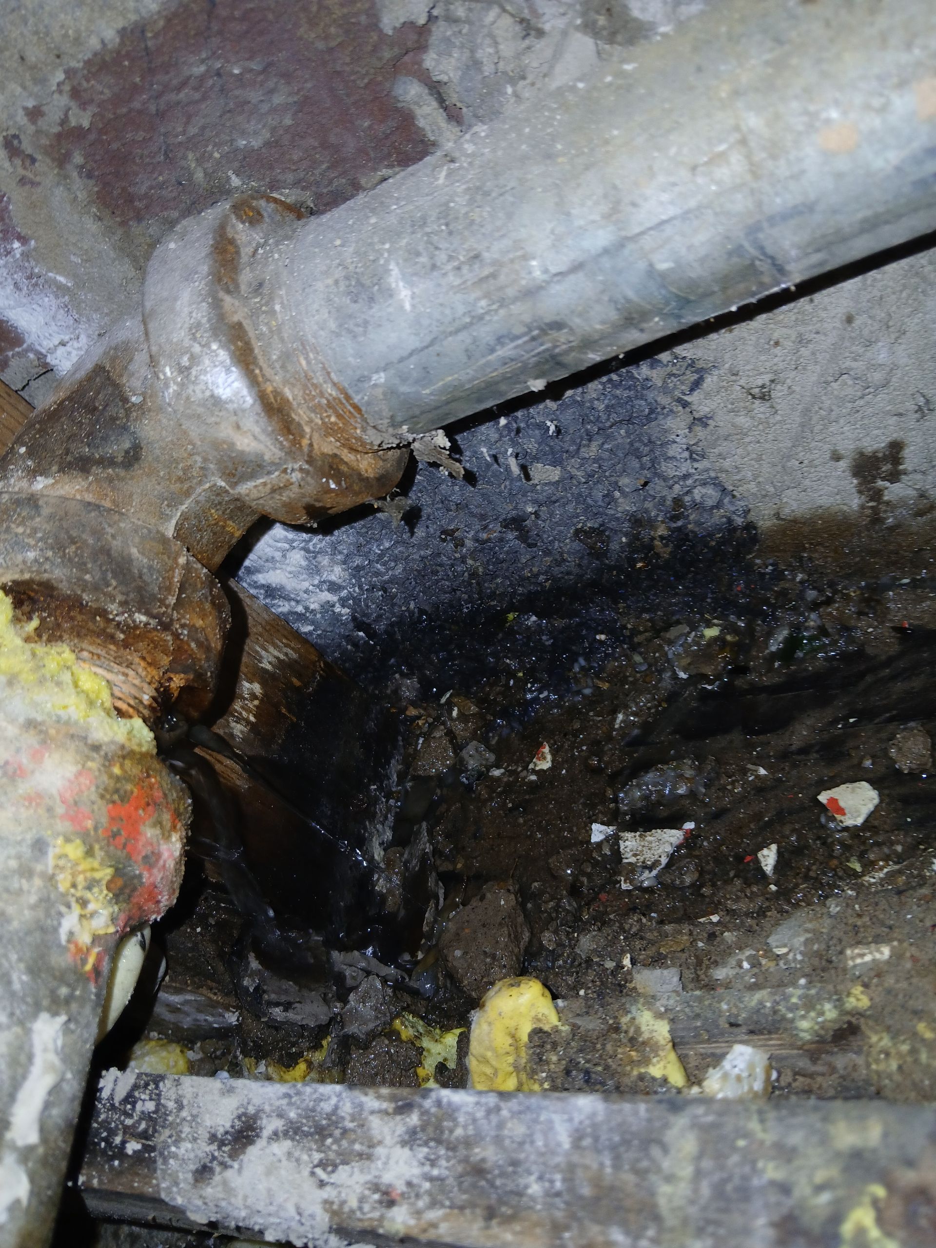 Corroded metal pipe joint with dark sludge and rust in a grimy drainage area