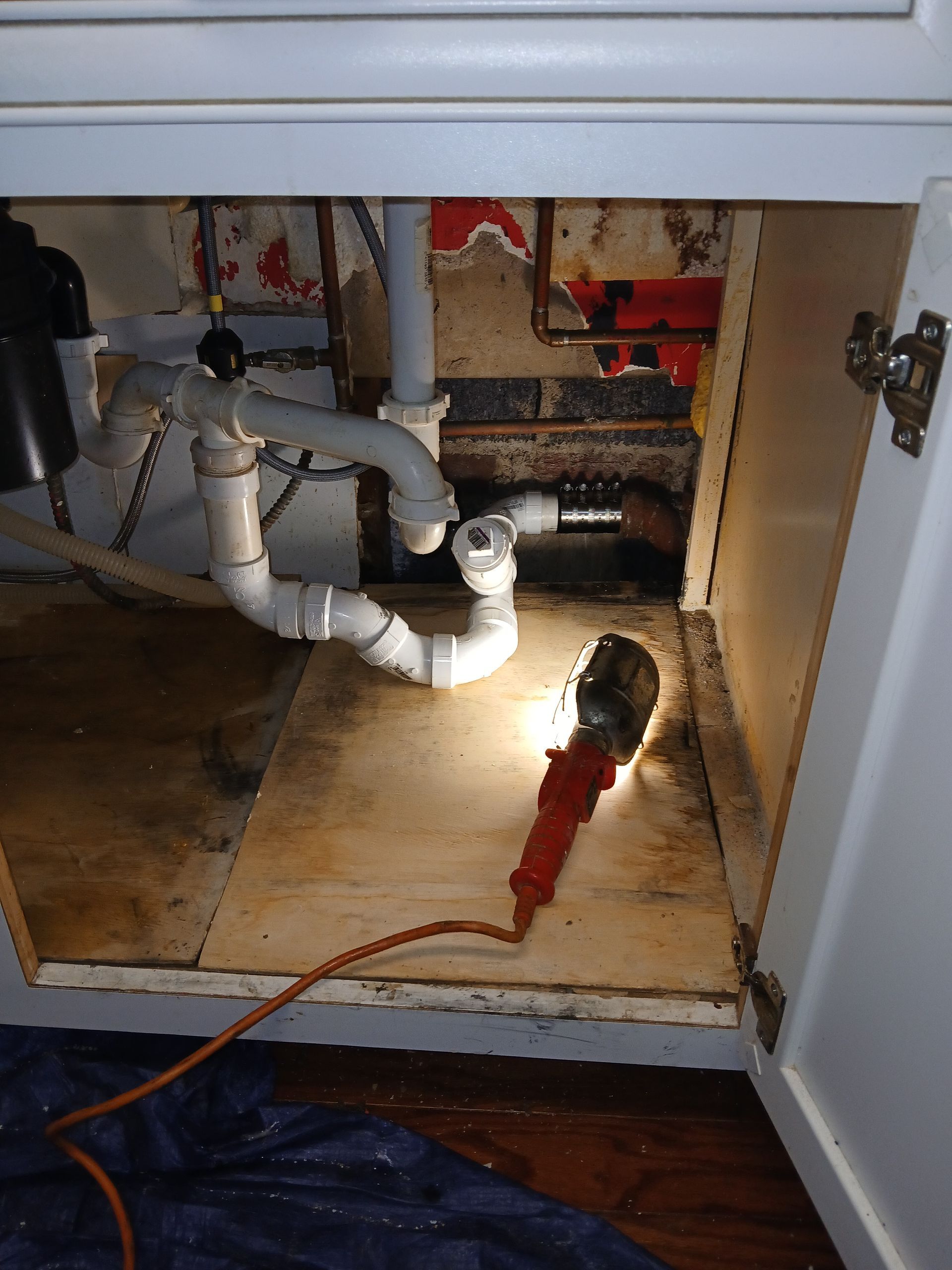 Under-sink plumbing with new exposed white PVC pipes, open cabinet, and a red flashlight on the floor