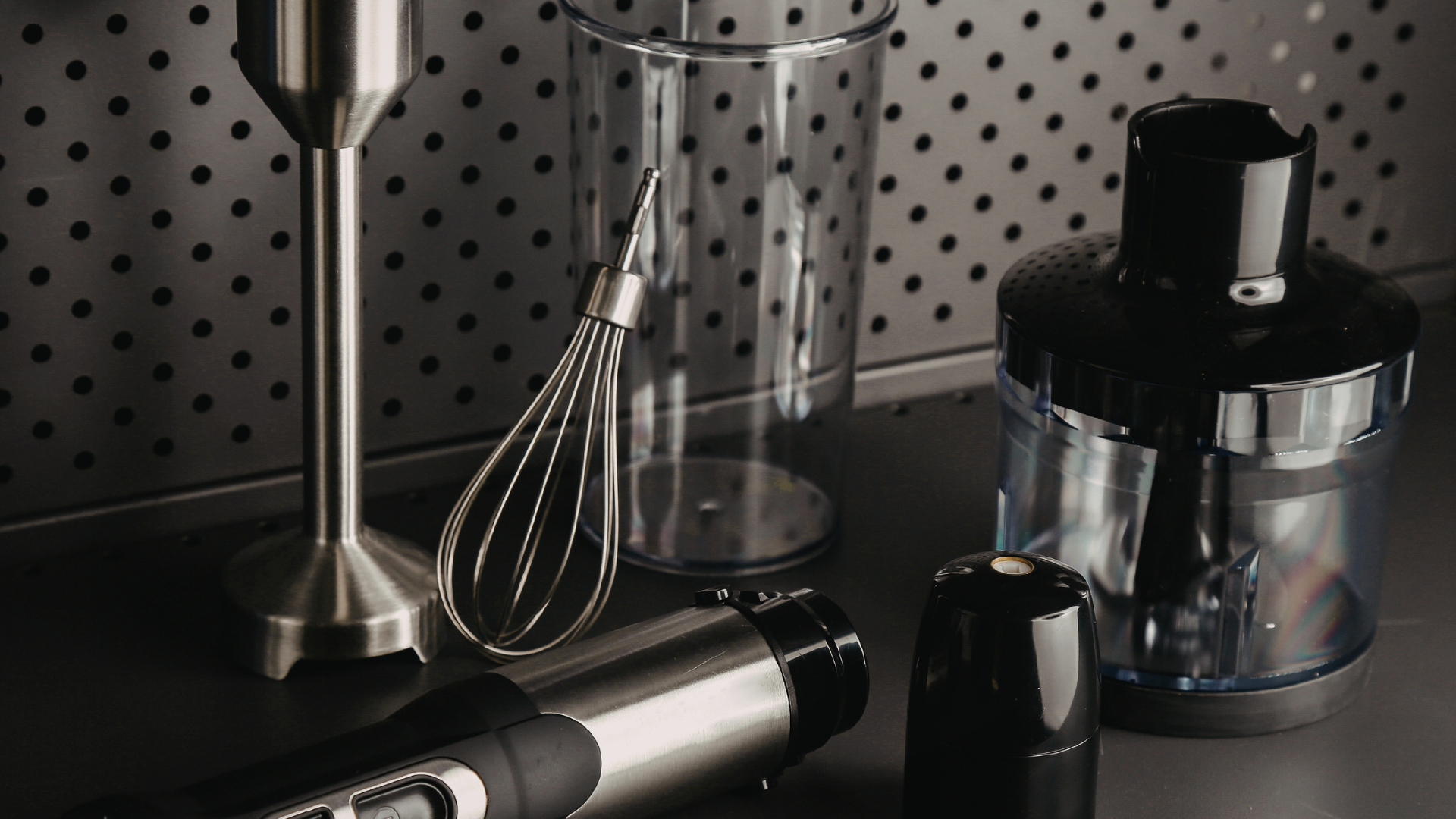 An immersion blender set with a metal whisk and blending container on a dark surface against a pegboard wall.