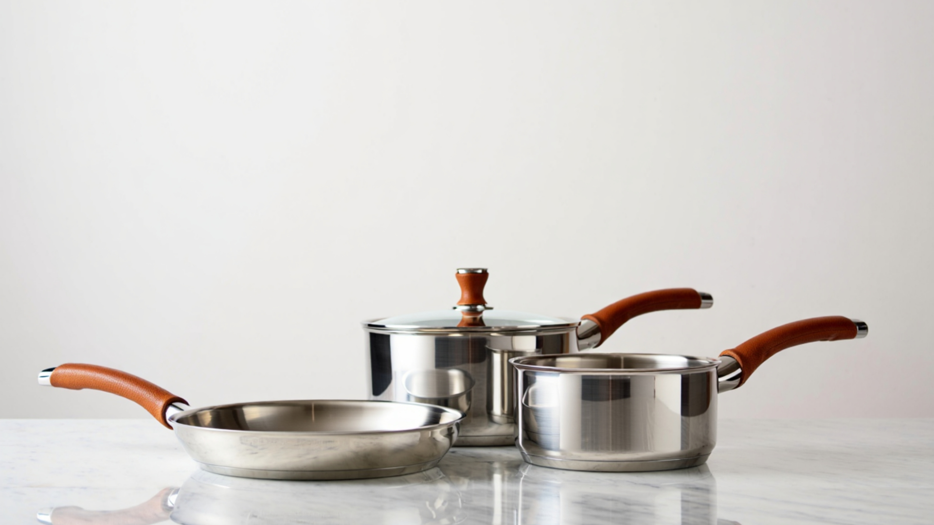 Three stainless steel kitchen pans with brown handles, including one with a lid, sitting on a white marble surface.