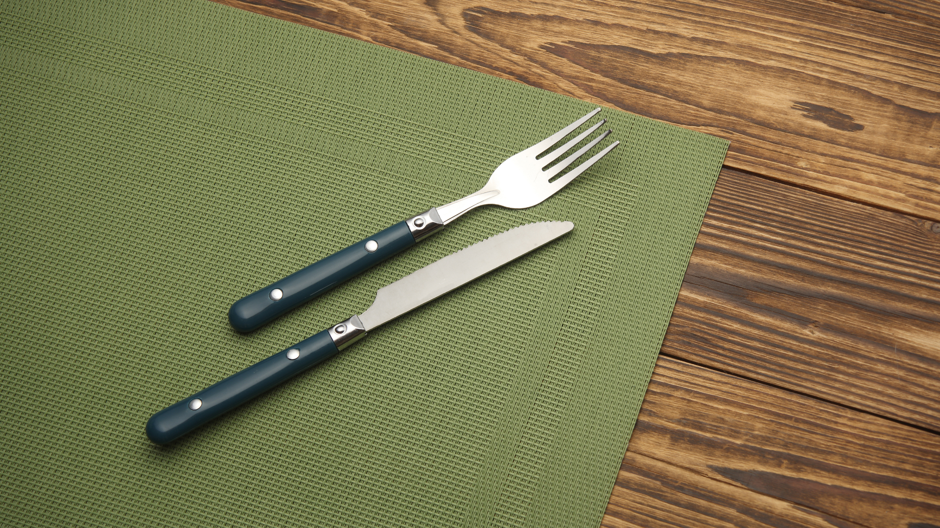A fork and a knife with dark handles placed diagonally on a green textured placemat on a wooden surface.