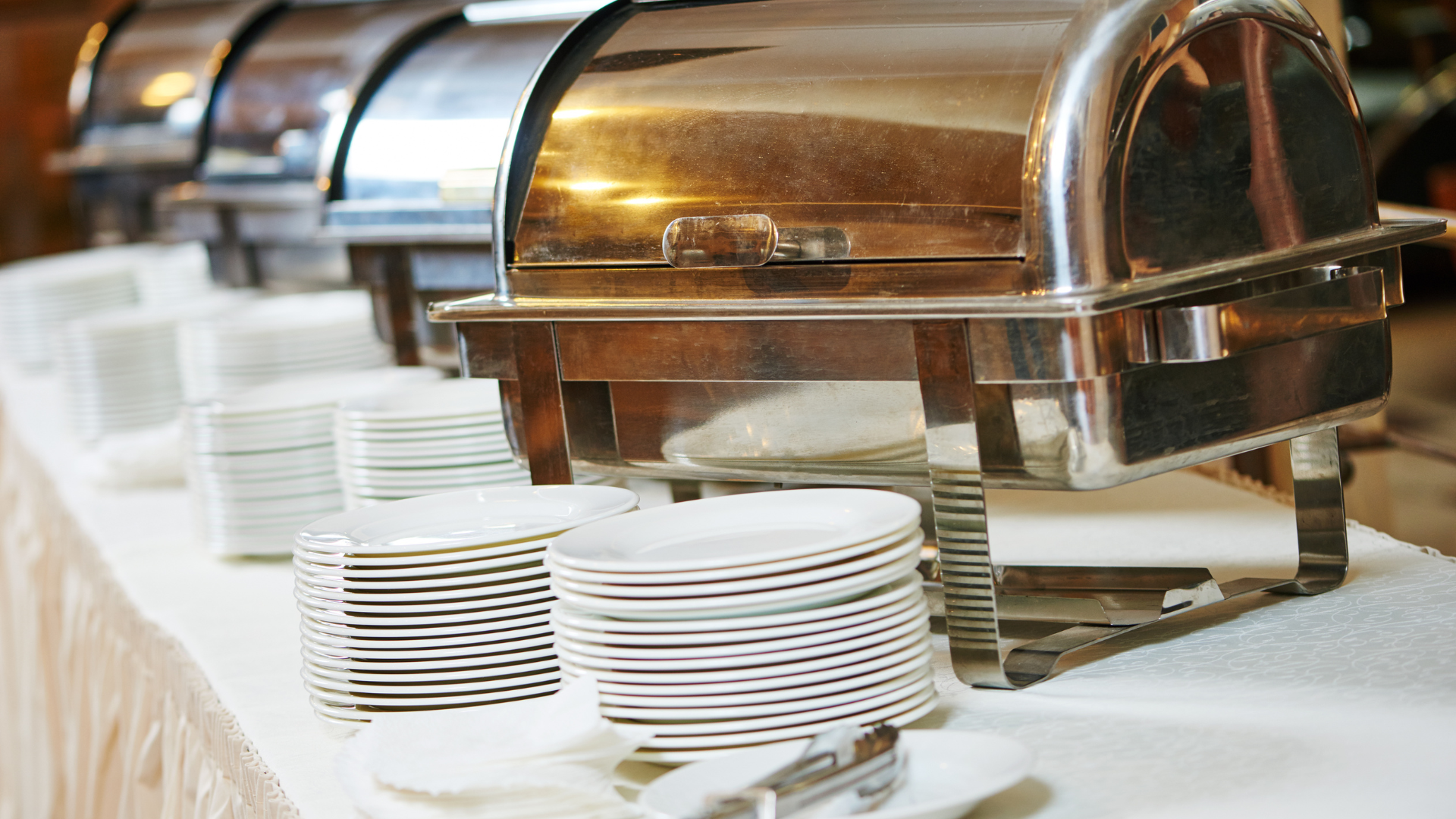 A buffet table with rows of metal chafing dishes and stacks of white dinner plates.