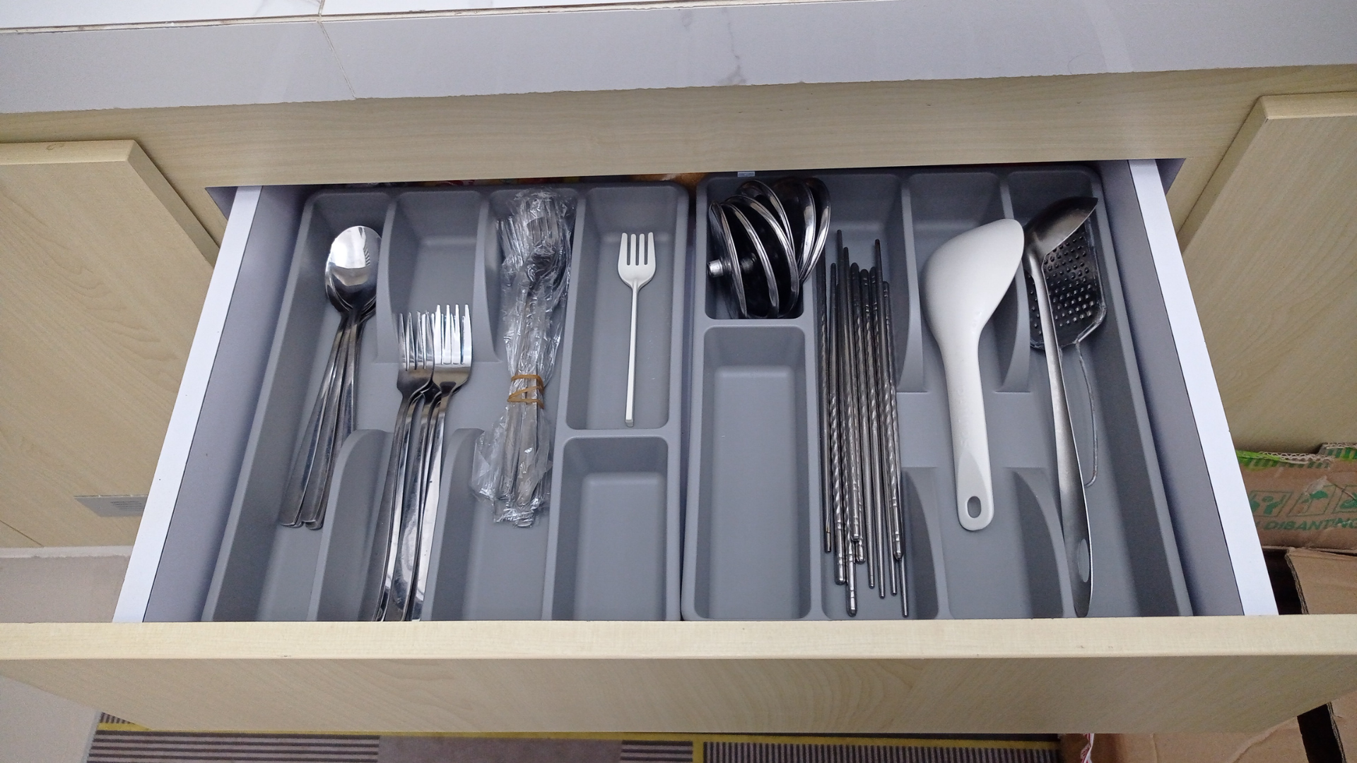A grey cutlery organizer in a kitchen drawer holds spoons, forks, chopsticks, and a large white serving spoon.