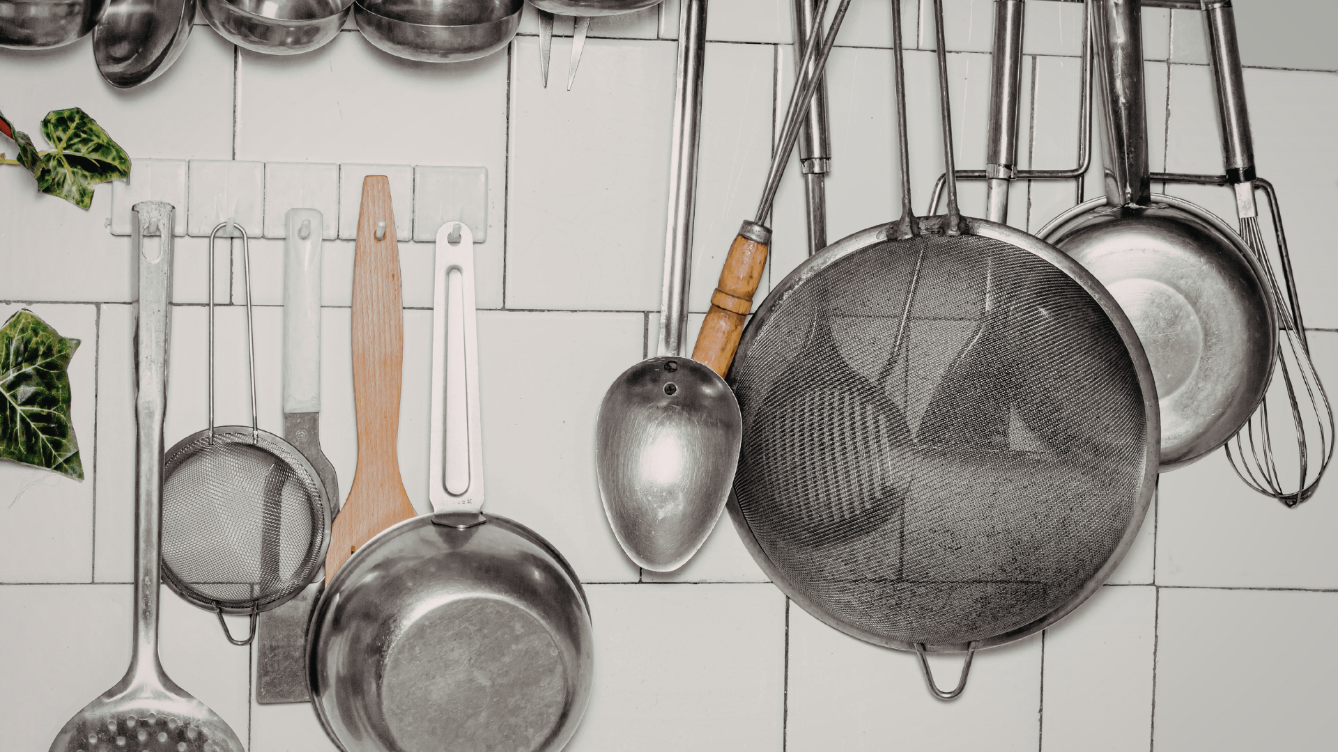 A variety of stainless steel kitchen utensils and strainers hang against a white tiled wall.