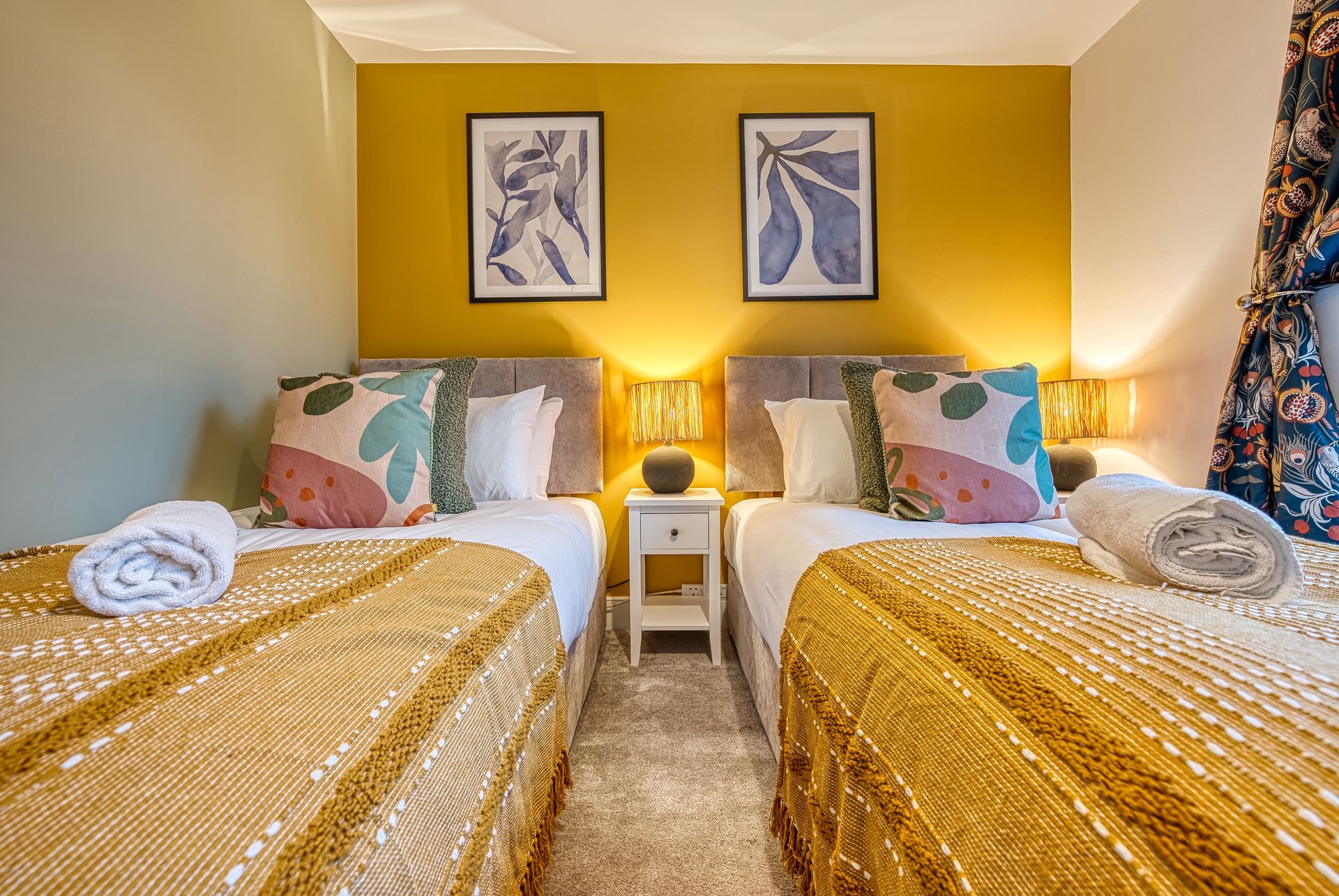 Twin beds in a bedroom with a yellow wall, artwork, and patterned throw blankets.