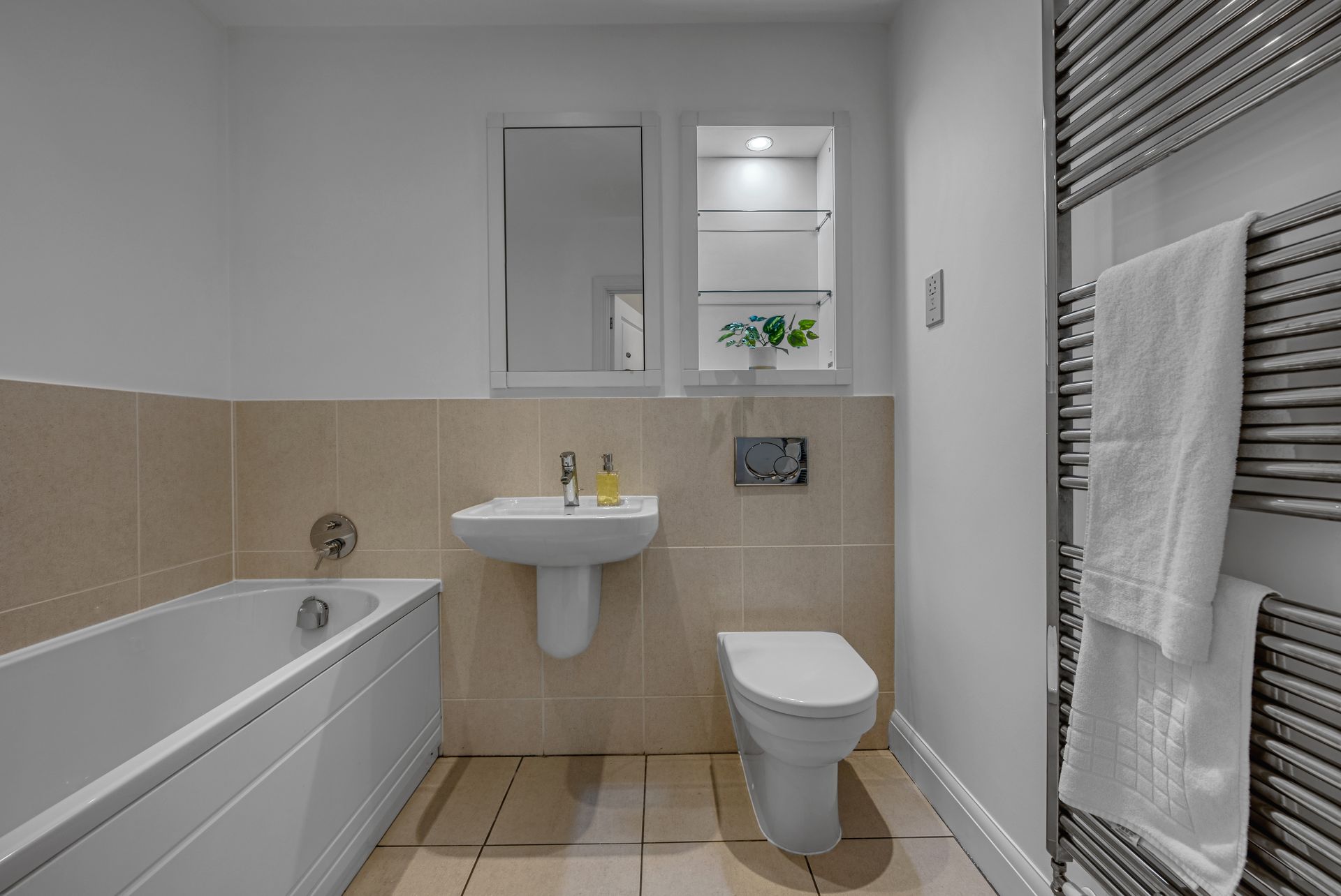 Bathroom with a white bathtub, toilet, sink, and heated towel rack. Beige tiles and walls.