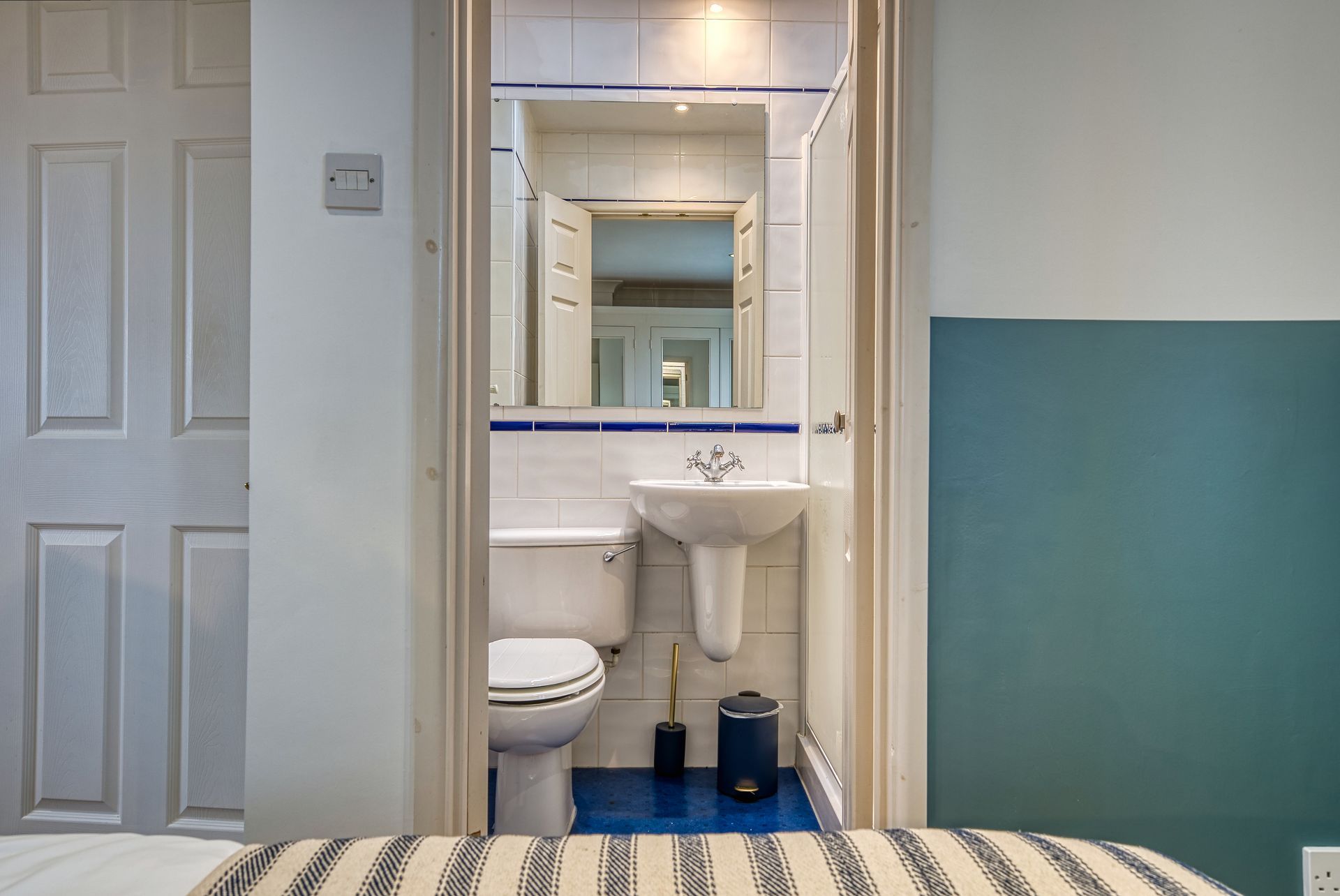 Bathroom: white toilet, sink, mirror. Blue tile floor, doorframe in bedroom.