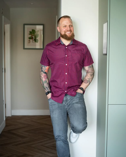 Man with tattoos smiles, leans against wall in hallway, wearing purple shirt and jeans.
