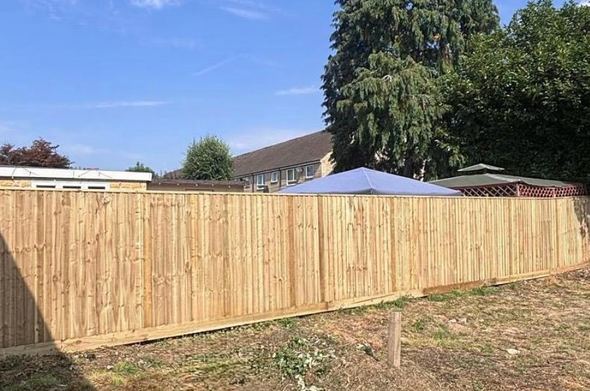 Wooden fence in front of a house, with green trees and a blue-roofed structure in the background.