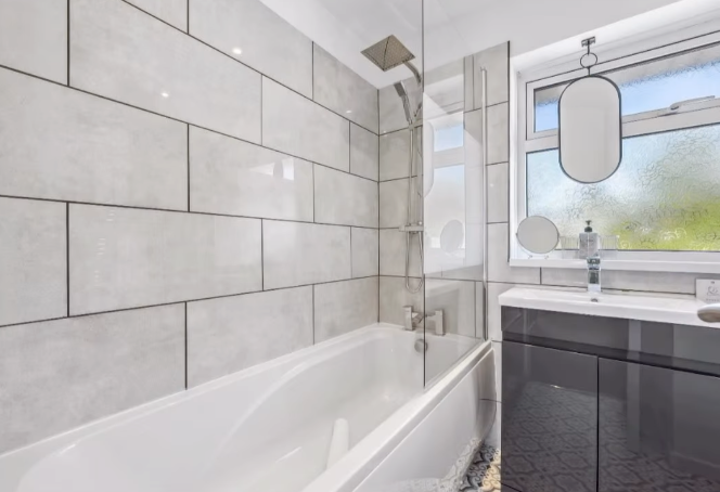 Bathroom with white tub, grey tiled walls, glass shower screen, and dark gray vanity.