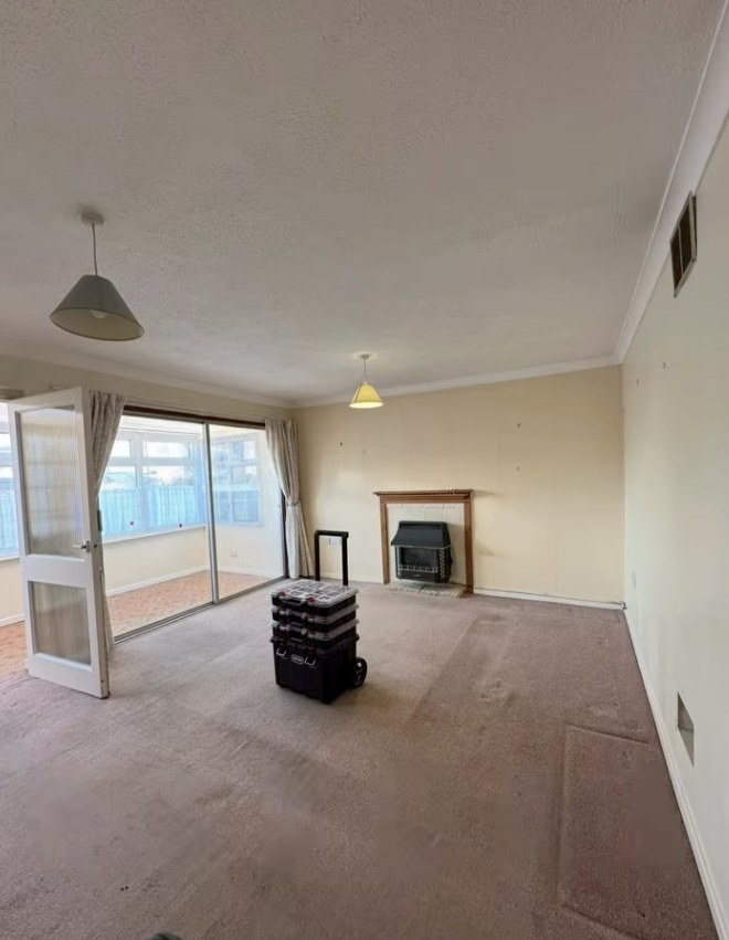 Empty living room with beige walls, carpet, and a fireplace. Sliding glass doors lead to a porch.