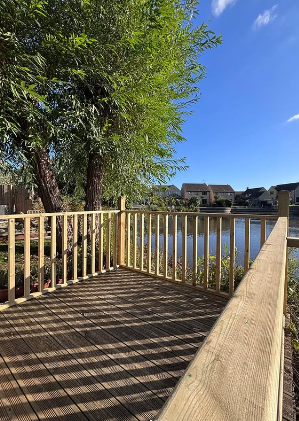 Wooden deck and railing overlooking a lake, with trees and houses in the background under a blue sky.