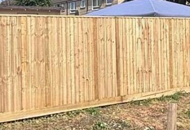 Wooden fence, light brown, vertical planks, in front of a building with a blue tarp.