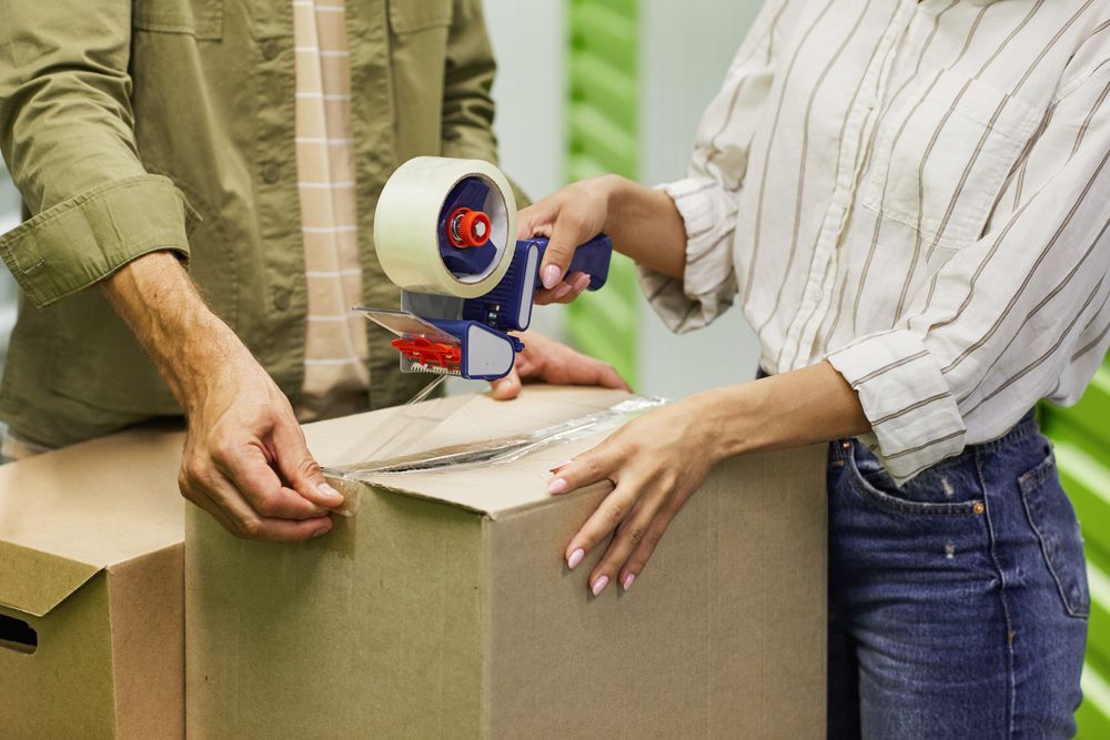 A Man And A Woman Are Wrapping A Cardboard Box With Tape — Ahern's Removals In Tamworth, NSW