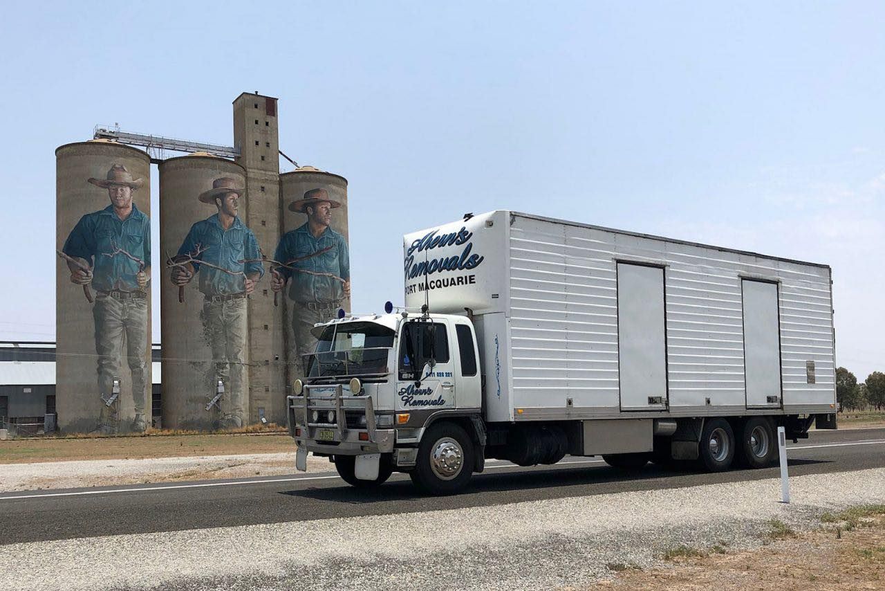 A White Truck Is Driving Down A Road In Front Of Silos — Ahern's Removals In South West Rocks, NSW