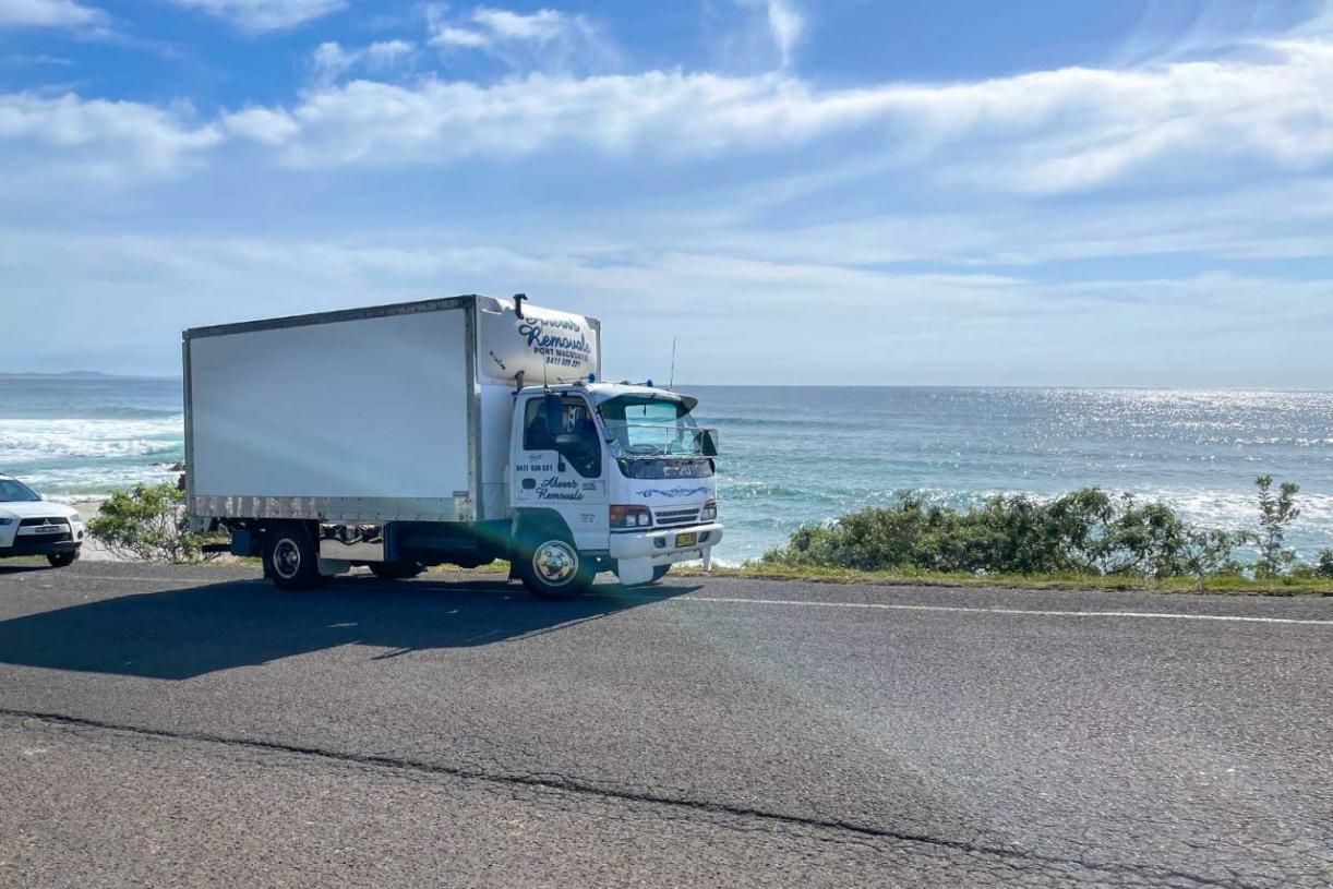 A White Truck Is Driving Down A Road Next To The Ocean — Ahern's Removals In Laurieton, NSW