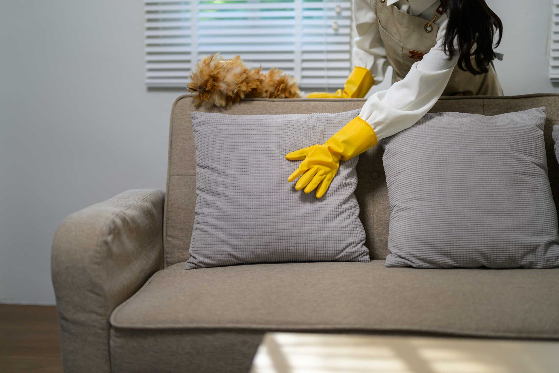 A woman wearing yellow gloves adjusting gray cushions on a beige sofa with a feather duster nearby