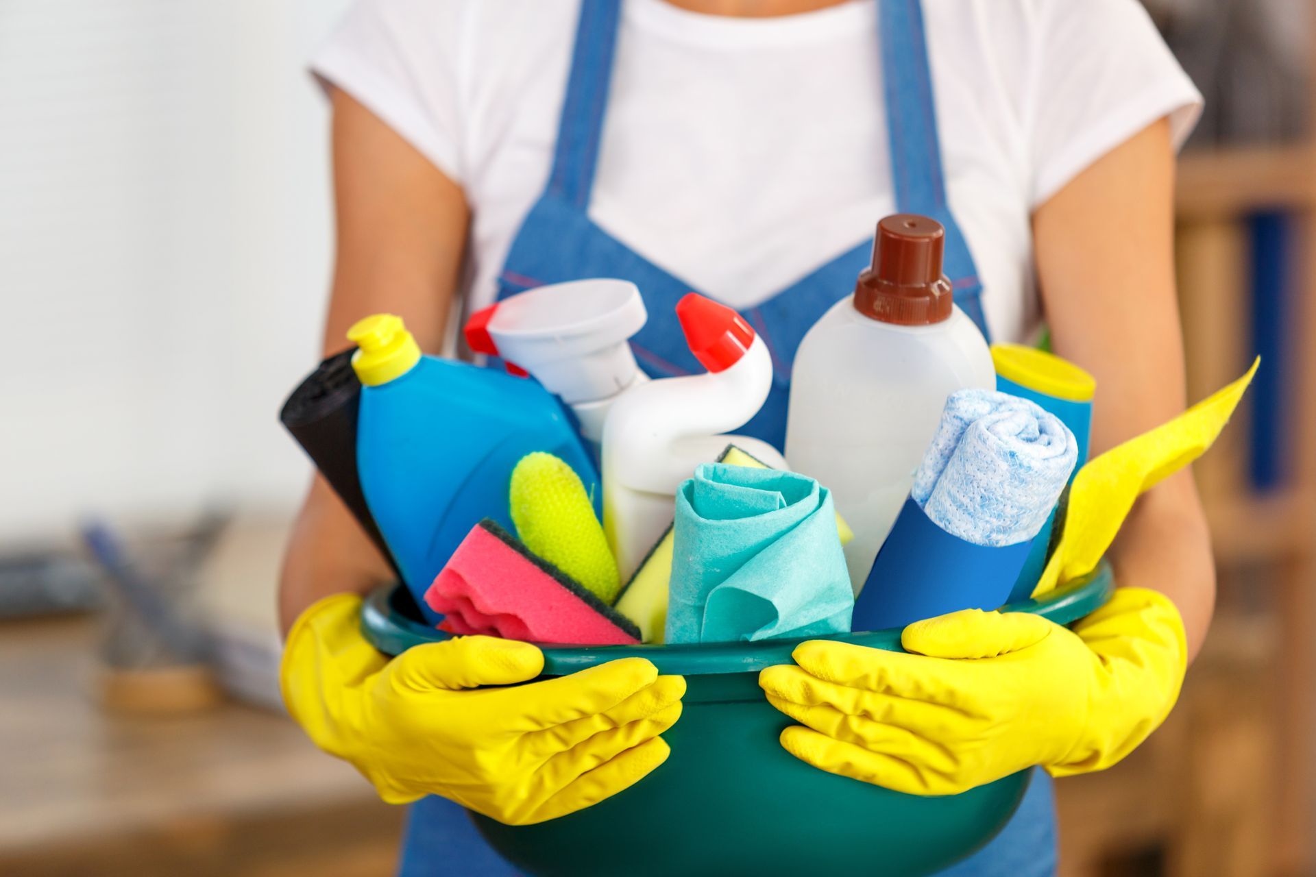 Cleaning supplies in bucket on kitchen floor with water spill and person in background. Person in yellow gloves holding a blue cleaning caddy with colorful spray bottles and supplies