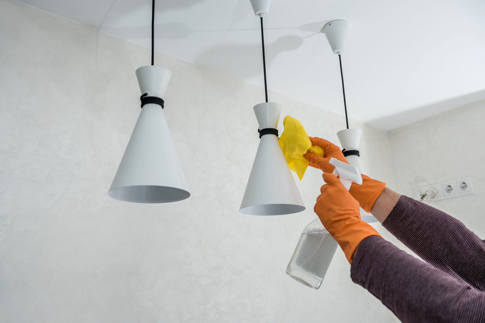 A person wearing orange gloves cleaning white pendant lights with a yellow cloth and spray bottle