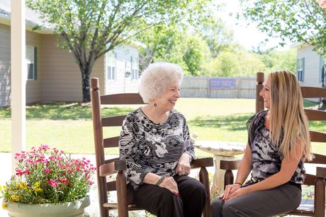 Two women smiling, sitting on rocking chairs on a porch, potted flowers nearby.
