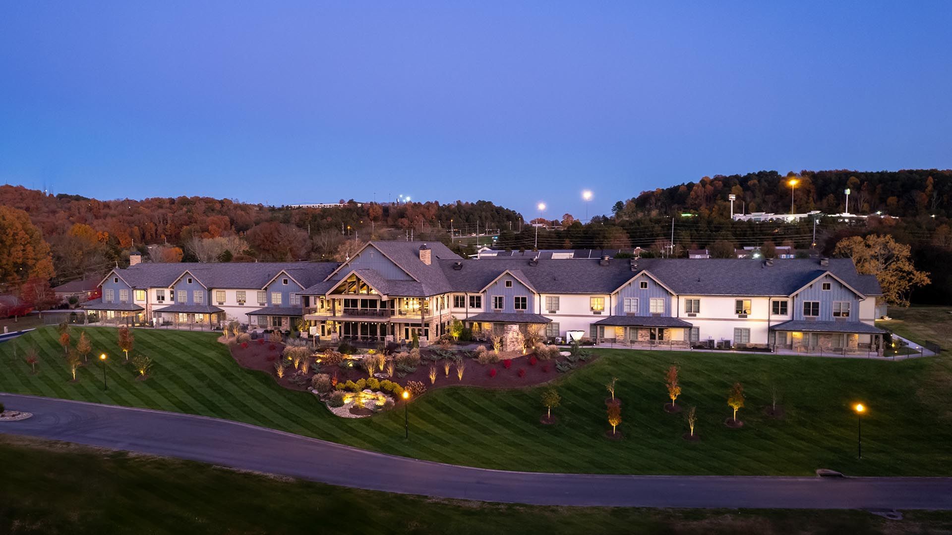 Large, multi-level lodge-style building on a grassy hill, with a pond, under a dusky blue sky.